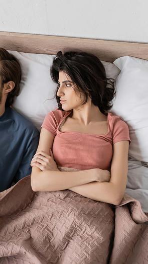 Een koppel ligt in bed. De man kijkt afwezig weg en de vrouw is ontevreden.