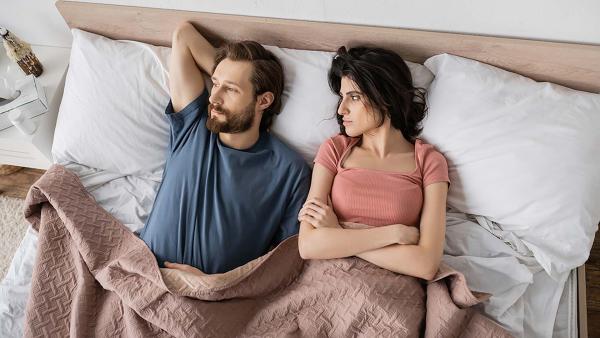 Een koppel ligt in bed. De man kijkt afwezig weg en de vrouw is ontevreden.