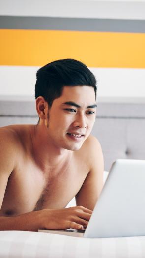 Man ligt zonder shirt op bed en bekijkt iets op zijn laptop