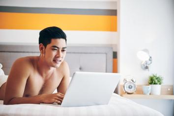 Man ligt zonder shirt op bed en bekijkt iets op zijn laptop