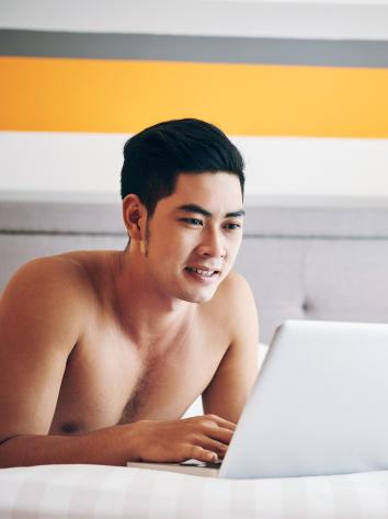 Man ligt zonder shirt op bed en bekijkt iets op zijn laptop