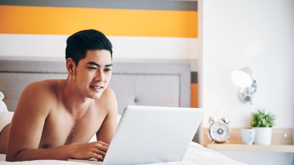 Man ligt zonder shirt op bed en bekijkt iets op zijn laptop
