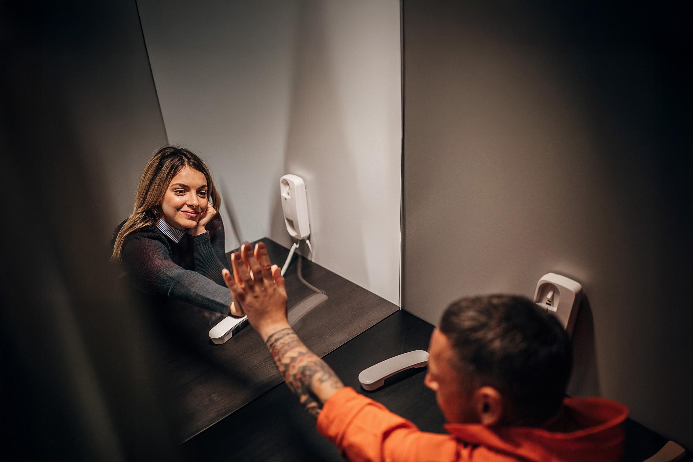 Vrouw op bezoek in gevangenis met hand tegen glazen wand tegen hand van gevangene. Beide met telefoon aan oor. 