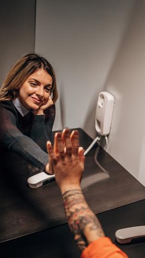 Vrouw op bezoek in gevangenis met hand tegen glazen wand tegen hand van gevangene. Beide met telefoon aan oor. 