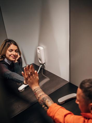 Vrouw op bezoek in gevangenis met hand tegen glazen wand tegen hand van gevangene. Beide met telefoon aan oor. 