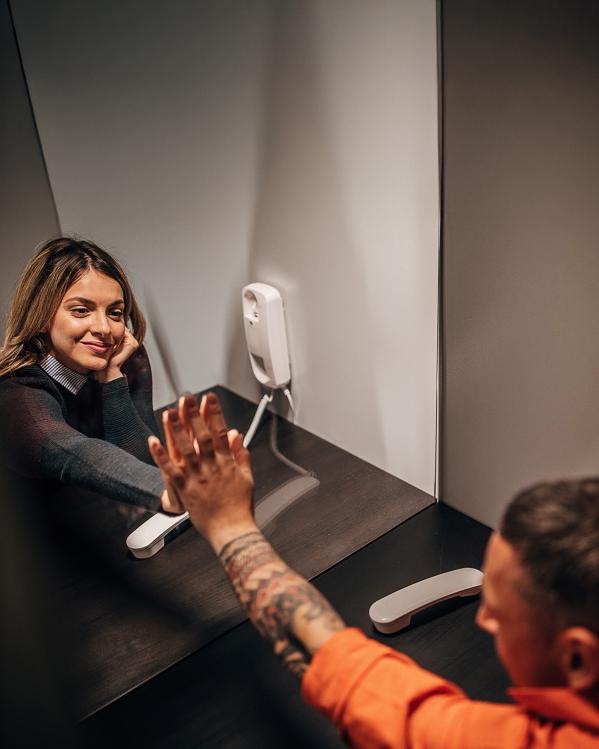 Vrouw op bezoek in gevangenis met hand tegen glazen wand tegen hand van gevangene. Beide met telefoon aan oor.