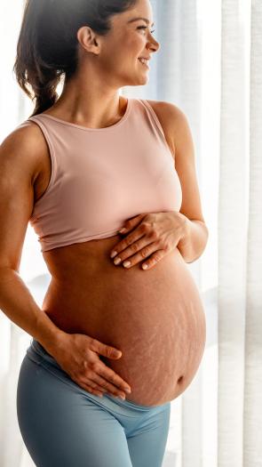 Zwangere vrouw kijkt lachend weg met handen op buik met zichtbare striae