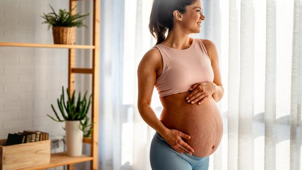 Zwangere vrouw kijkt lachend weg met handen op buik met zichtbare striae