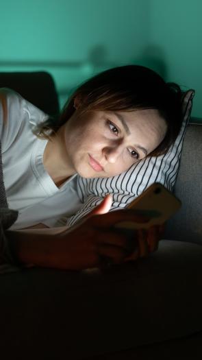 Vrouw ligt onder kleedje op de bank in donkere kamer en kijkt naar telefoon waar blauw licht uit schijnt.