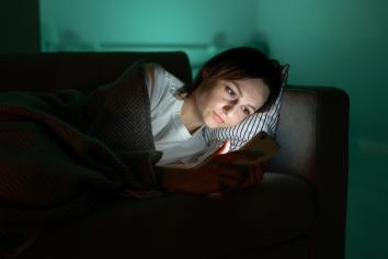 Vrouw ligt onder kleedje op de bank in donkere kamer en kijkt naar telefoon waar blauw licht uit schijnt.