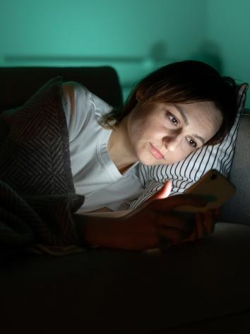 Vrouw ligt onder kleedje op de bank in donkere kamer en kijkt naar telefoon waar blauw licht uit schijnt.
