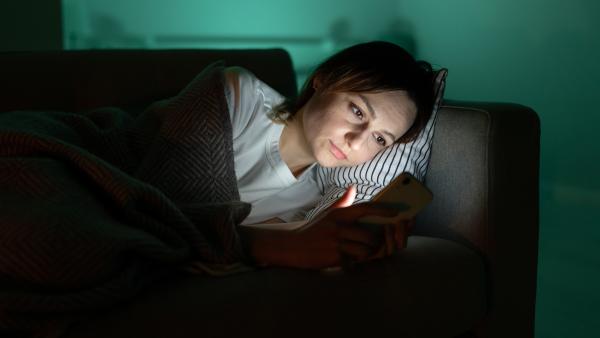 Vrouw ligt onder kleedje op de bank in donkere kamer en kijkt naar telefoon waar blauw licht uit schijnt.