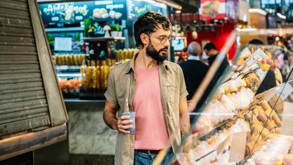 Man met bril en blauwe smoothie kijkt naar vitrine met soorten gebak