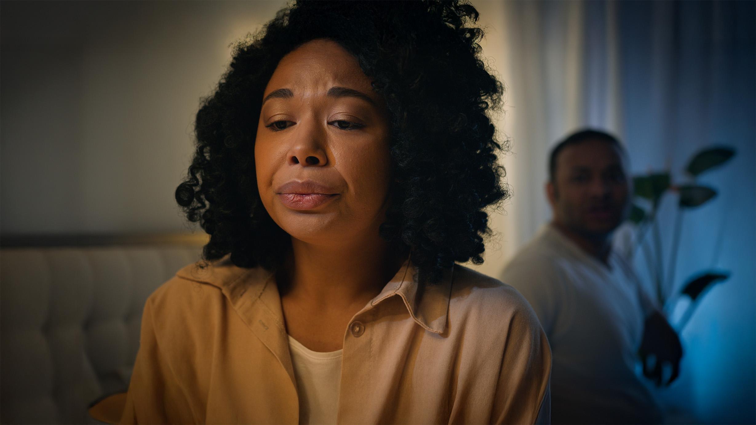Vrouw staart emotioneel voor zich uit terwijl een man met armen over elkaar heen naar haar rug kijkt