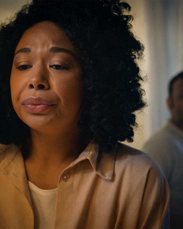 Vrouw staart emotioneel voor zich uit terwijl een man met armen over elkaar heen naar haar rug kijkt