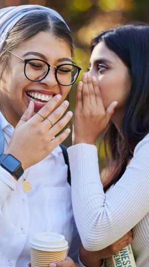 Een vrouw fluistert iets in het oor van een andere vrouw, die hier om moet lachen. 
