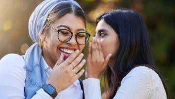 Een vrouw fluistert iets in het oor van een andere vrouw, die hier om moet lachen. 