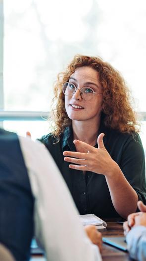 Vier personen aan tafel. Kijken allemaal naar een vrouw met rode krullen en een bril die het woord heeft