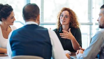 Vier personen aan tafel. Kijken allemaal naar een vrouw met rode krullen en een bril die het woord heeft