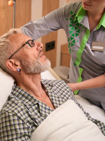 Man met bril en baard in ziekenhuis bed terwijl verpleegkundige zijn kussen recht legt