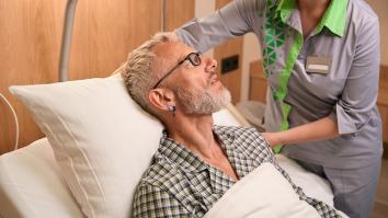 Man met bril en baard in ziekenhuis bed terwijl verpleegkundige zijn kussen recht legt