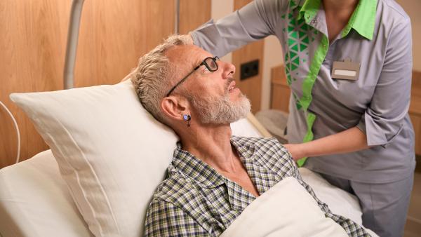Man met bril en baard in ziekenhuis bed terwijl verpleegkundige zijn kussen recht legt