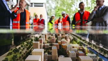 En maquette van een nieuwbouwwijk met boompjes en water. Daaromheen staan mensen in oranje en gele hesjes.
