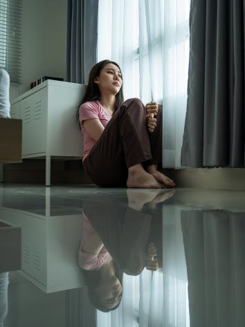 Vrouw zit op haar slaapkamervloer met knieën omhoog en kijkt bedroefd naar buiten langs het gordijn