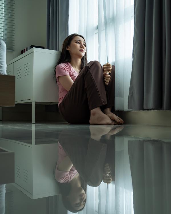 Vrouw zit op haar slaapkamervloer met knieën omhoog en kijkt bedroefd naar buiten langs het gordijn