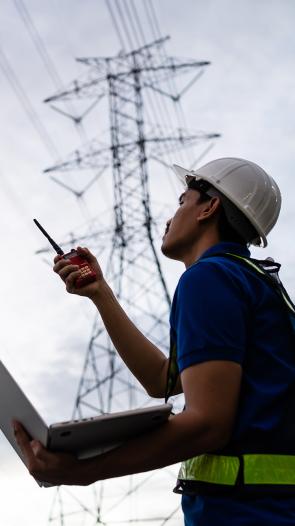een man met bouwvakkershelm op en laptop in hand kijkt naar elektriciteitsmasten