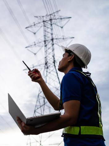 een man met bouwvakkershelm op en laptop in hand kijkt naar elektriciteitsmasten