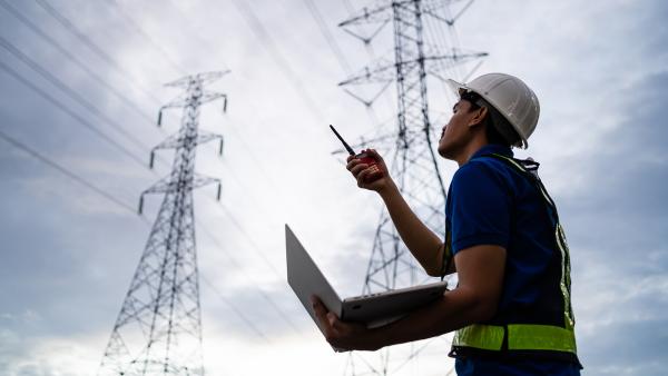 een man met bouwvakkershelm op en laptop in hand kijkt naar elektriciteitsmasten