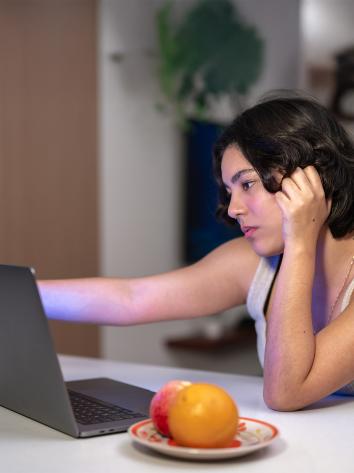 Vrouw zit aan tafel achter laptop. Houdt laptopscherm vast en kijkt bedroefd. 