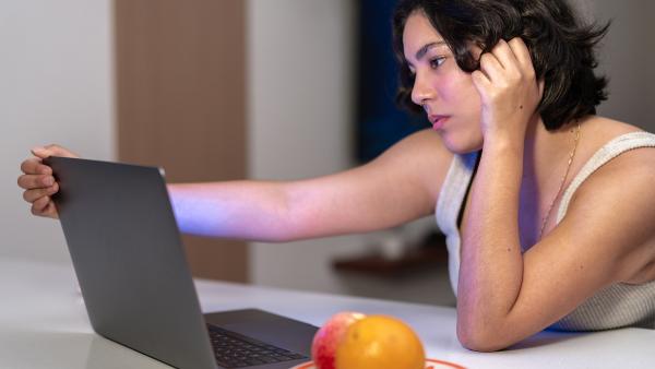 Vrouw zit aan tafel achter laptop. Houdt laptopscherm vast en kijkt bedroefd. 