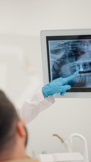 Tandarts met medische handschoen wijst naar röntgenfoto van gebit met man in tandartsstoel ernaast