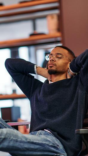 Jongen zit met zijn voeten op zijn bureau, met daarop meerdere schermen. Hij heeft zijn ogen dicht en handen achter zijn hoofd.