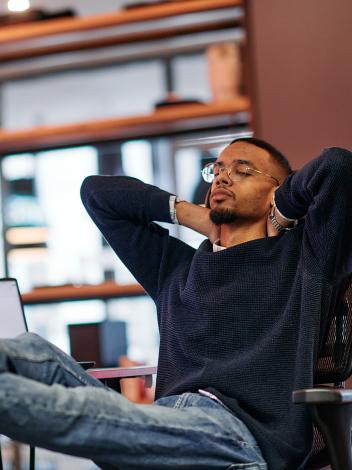 Jongen zit met zijn voeten op zijn bureau, met daarop meerdere schermen. Hij heeft zijn ogen dicht en handen achter zijn hoofd.