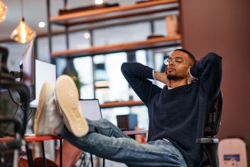 Jongen zit met zijn voeten op zijn bureau, met daarop meerdere schermen. Hij heeft zijn ogen dicht en handen achter zijn hoofd.