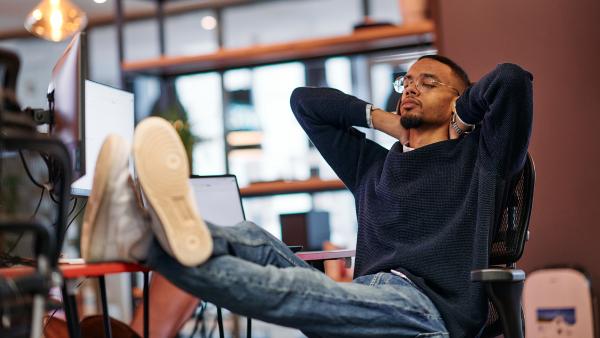 Jongen zit met zijn voeten op zijn bureau, met daarop meerdere schermen. Hij heeft zijn ogen dicht en handen achter zijn hoofd.
