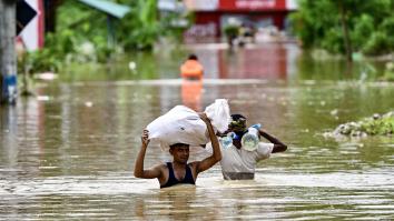 Twee mannen lopen tot hun borst in het water over straat met een zak eten en flessen drinkwater boven hun hoofd