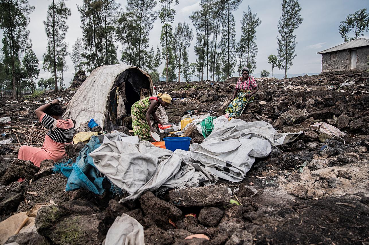 Drie vrouwen zitten voor een tent tussen de restanten van een afgebrand vluchtelingenkamp. Een vrouw doet de afwas en de ander vlecht haar haar.