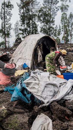 Drie vrouwen zitten voor een tent tussen de restanten van een afgebrand vluchtelingenkamp. Een vrouw doet de afwas en de ander vlecht haar haar. 