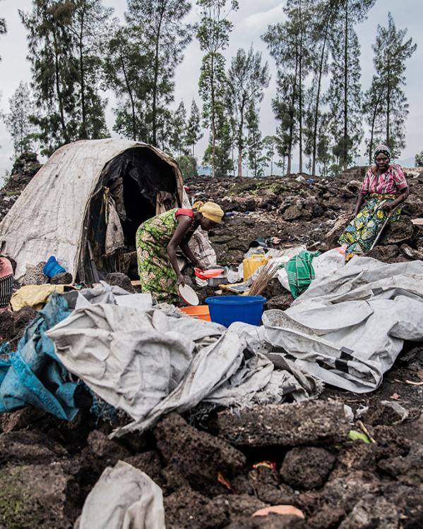 Drie vrouwen zitten voor een tent tussen de restanten van een afgebrand vluchtelingenkamp. Een vrouw doet de afwas en de ander vlecht haar haar. 