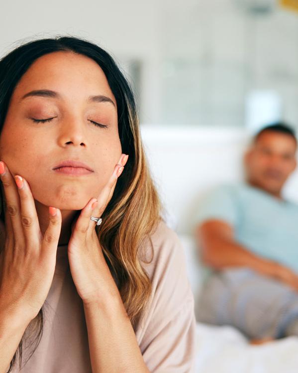Vrouw zit op bed met hoofd in handen en ogen dichtgeknepen. Gespannen gezichtsuitdrukking. Onscherp zit een man op de achtergrond.