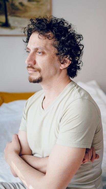 Een man zit op de rand van een bed met zijn armen over elkaar, en kijkt somber naar beneden.