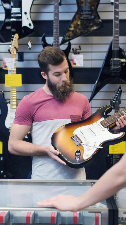 Een foto van een man in een muziekwinkel die een elektrische gitaar bekijkt, terwijl de winkelier wacht aan de toonbank. Achter hem hangen verschillende gitaren aan de muur.