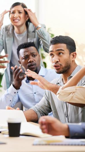 Groep mensen zit aan een tafel en discussieert met veel handgebaren, zoals wijzen naar elkaar en handen tegen het hoofd houden