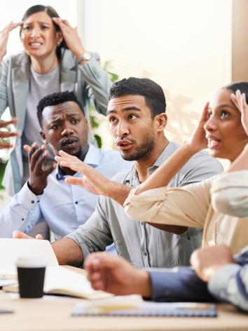Groep mensen zit aan een tafel en discussieert met veel handgebaren, zoals wijzen naar elkaar en handen tegen het hoofd houden