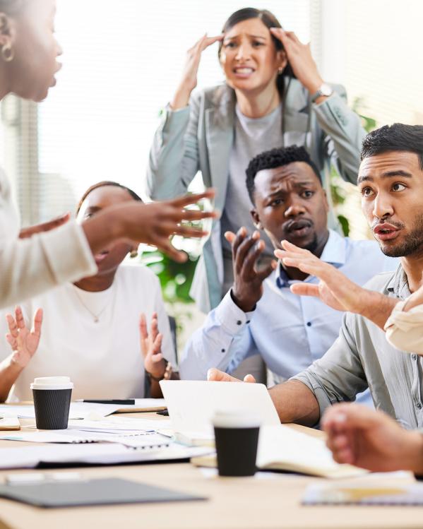 Groep mensen zit aan een tafel en discussieert met veel handgebaren, zoals wijzen naar elkaar en handen tegen het hoofd houden