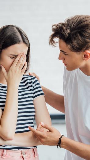 Een foto van een jonge man die een verdrietige jonge vrouw troost door een hand op haar schouder te leggen en haar aan te kijken.